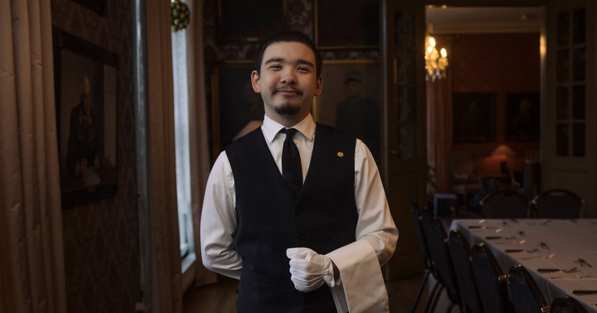 En smilende servitør i formell uniform står i et elegant, historisk selskapsrom med dekkede bord og dempet belysning.