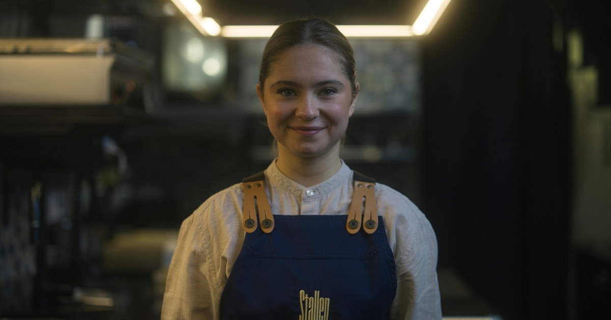 Person i forkle står på et kjøkken og smiler mot kamera.