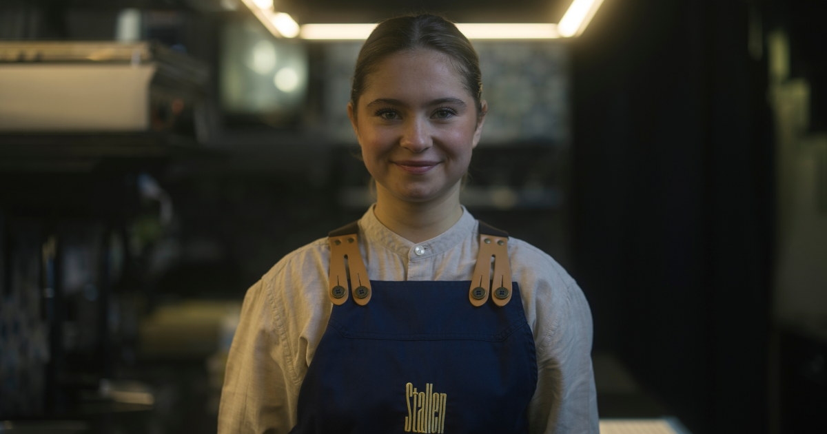 Person i forkle står på et kjøkken og smiler mot kamera.